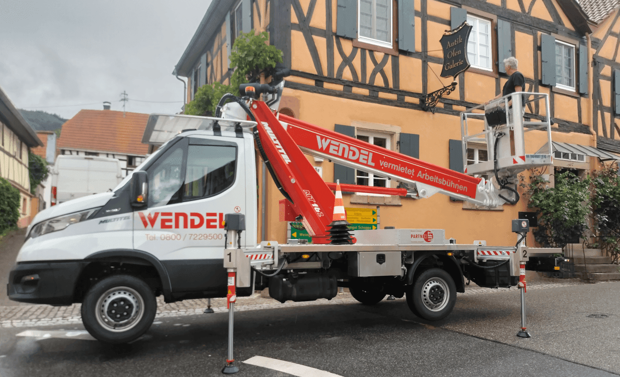 Das Bild zeigt einen Wagen mit einem kleinen Kran vor einem orangefarbenen Fachwerkhaus, dem ehemaligen Standort der Antik Ofen Galerie. Das Firmenschild der Antik Ofen Galerie wird gerade abmontiert.